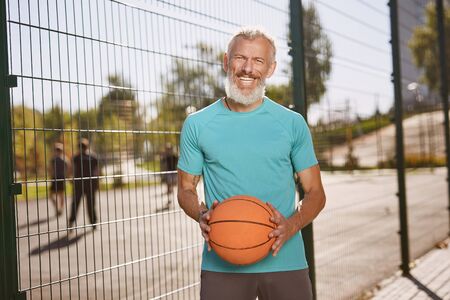 Basketball player. Happy mature man in sportswear holding basketball ball and smiling at camera while standing at outdoor basketball courtの写真素材