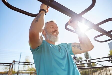 Strong and healthy people. Happy bearded senior man pulling up on horizontal bar and smiling while training outdoors. He is working out at the stadium. Fitness, sport, workout, healthy lifestyleの写真素材