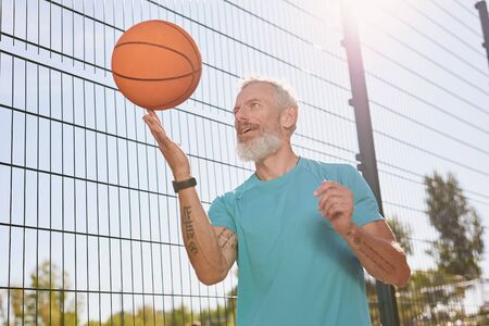 Playing basketball outdoors. Happy elderly man in sportswear spinning a basketball ball on his finger while standing at outdoor basketball courtの写真素材