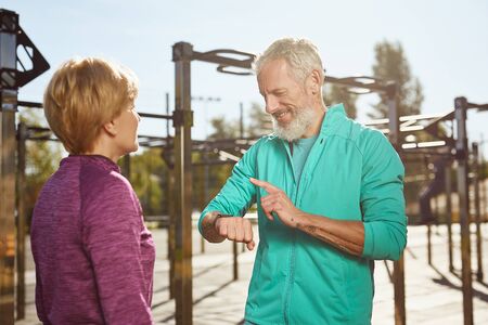 Checking fitness results. Happy senior man in sportswear looking at smartwatch and smiling while exercising with his wife at outdoor gym. Active family coupleの写真素材