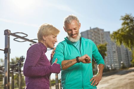 Great results. Happy mature family couple in sportswear looking at smartwatch and checking training results while standing outdoorsの写真素材