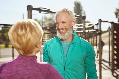 Active morning. Happy senior family couple is doing sport outdoors. Mature bearded man in sport wear talking with his wife while working out together at outdoor gymの写真素材