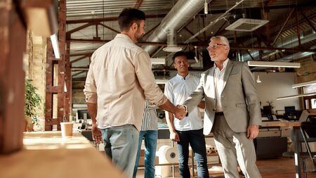 Welcome to our team! Two cheerful men shaking hands and smiling while standing with coworkers in the modern officeの写真素材