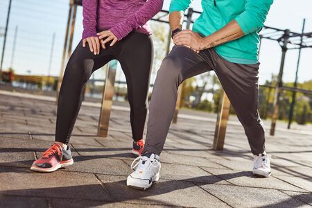Stretching legs. Cropped photo of a couple in sportswear warming up together at the stadium. Man and woman doing stretching exercises outdoorsの写真素材