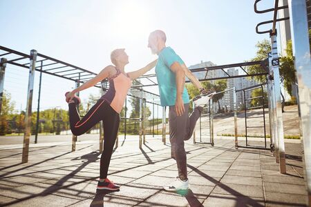 Happy mature man and woman in sportswear warming up together at the stadium in the morning. They stretching legs, looking at each other and smiling. Active couple doing sport. Healthy lifestyleの写真素材