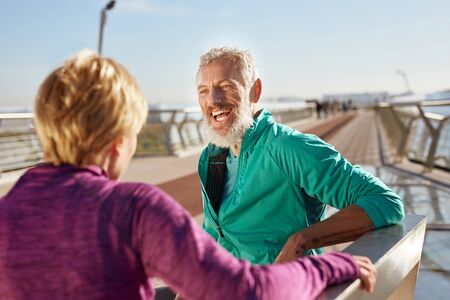 After morning run. Cheerful mature bearded man in sportswear talking with his wife and smiling while standing on the bridge in the morningの写真素材