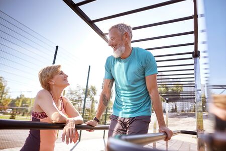 Happy mature bearded man in a great shape doing push ups on parallel bars and talking with his smiling wife. Active and healthy senior couple exercising in the morningの写真素材