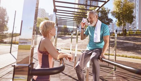 Tired mature couple resting after morning workout outdoors. They are standing near parallel bars and discussing something. Active senior couple doing sport together. Healthy lifestyle, sport, workoutの写真素材