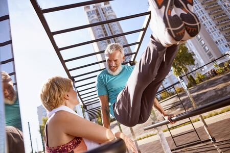 No age limit. Smiling mature man in great shape doing push ups on parallel bars and talking with his wife. Healthy mature couple doing sport together in the morning outdoors. Healthy active lifestyleの写真素材