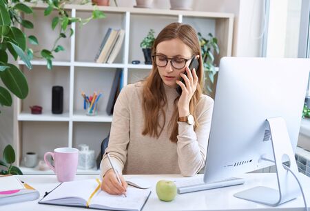 Young busy businesswoman, attractive female employee in glasses sitting at desk, talking by phone and making some notes while working remotely at homeの写真素材