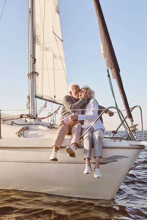 Happy senior couple sitting on the side of sail boat or yacht deck floating in sea. Man kissing woman, enjoying the viewの写真素材