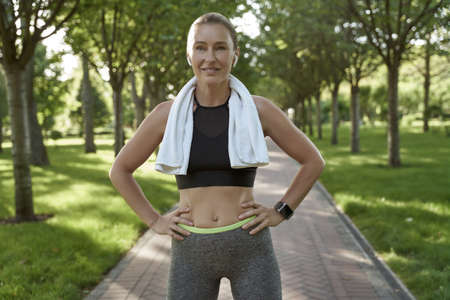 Beautiful athletic woman in sportswear and wireless headphones with towel on her shoulders looking at camera and smiling while exercising outdoorsの写真素材