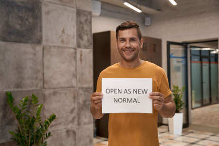 Young happy man, office worker showing paper with text OPEN AS NEW NORMAL at camera and smiling while standing in the modern coworking spaceの写真素材
