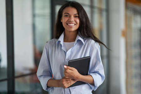 Confident business woman at work. Portrait of a young cheerful female employee holding digital tablet, looking at camera and smiling while standing in the modern officeの写真素材