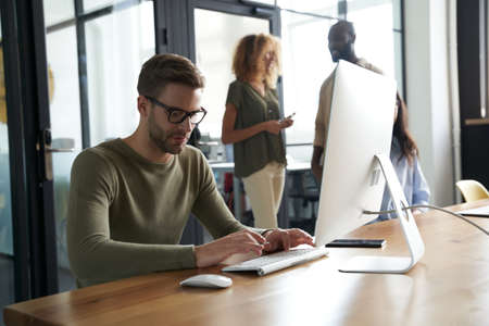 Young caucasian man, office worker typing something on computer while sitting at his workplace in the modern office with colleagues on the backgroundの写真素材