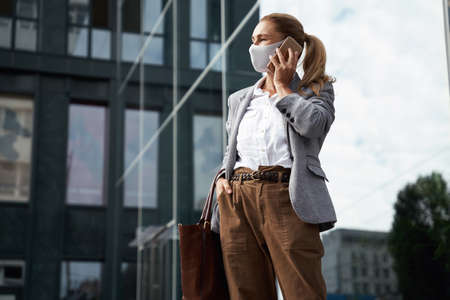 Working safely during coronavirus. Confident business woman wearing protective face mask talking on mobile phone while standing against office building outdoorsの写真素材