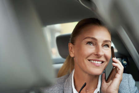 Good talk. Portrait of successful attractive business woman talking by mobile phone and smiling while sitting on back seat in the carの写真素材