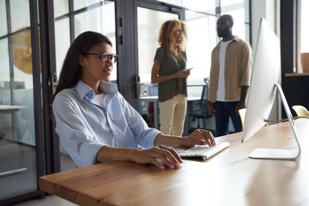 Young mixed race woman, female office worker working on computer while sitting at her workplace in the modern office, colleagues standing on the backgroundの写真素材