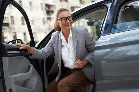 Portrait of attractive and happy business woman wearing eyeglasses getting out of her modern car and...