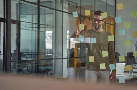 Planning new project. Young caucasian man, office worker wearing glasses writing something on sticky...