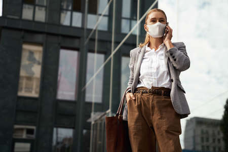 Business during covid 19. Confident business woman wearing protective face mask talking on mobile phone while standing against office building outdoorsの写真素材