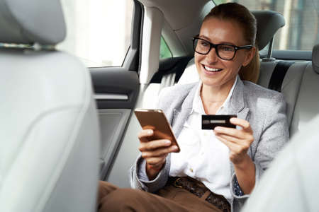 Cheerful business woman wearing eyeglasses using her smartphone and credit card to buy something and smiling while sitting on back seat in the carの写真素材