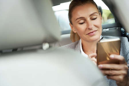 On the way to business meeting. Portrait of a beautiful and successful business woman using her smartphone and smiling while sitting on back seat in the carの写真素材