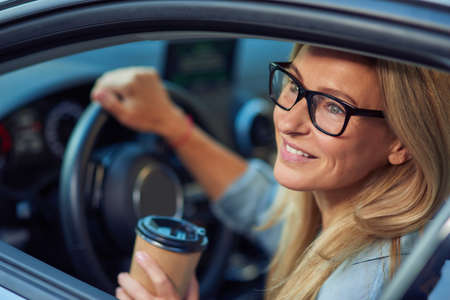 Smiling caucasian businesswoman wearing eyeglasses sitting behind steering wheel of her modern car and buying take away coffee in paper cupの写真素材