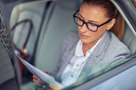 Concentrated middle aged businesswoman wearing eyeglasses reading documents while sitting on back seat in the car, preparing for a meetingの写真素材