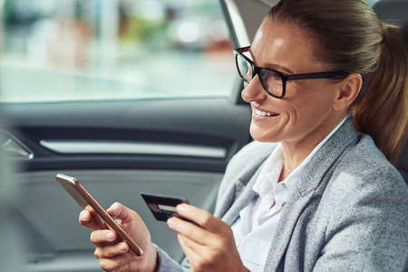 Pay online. Happy middle aged business woman using her smartphone and credit card to buy something while sitting on back seat in the carの写真素材