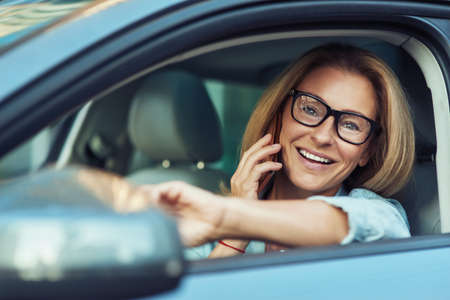 Portrait of successful happy business woman talking by mobile phone and smiling while driving a car, looking on the road through the windowの写真素材