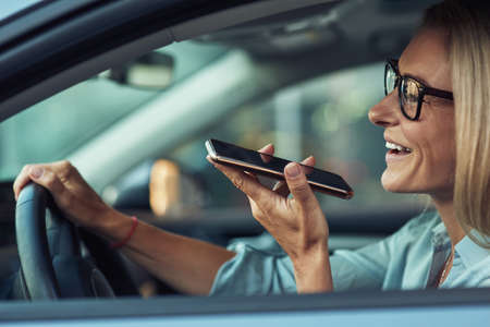 Side view of a smiling business woman using smartphone, recording voice message while driving a car