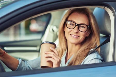 Portrait of a smiling mature woman wearing eyeglasses sitting behind steering wheel of her modern car, holding cup of take away coffee and looking at cameraの写真素材