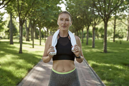 Workout in nature. Portrait of beautiful athletic woman in sportswear and wireless headphones with towel on her shoulders looking at camera while exercising outdoorsの写真素材