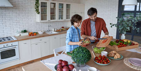 Healthy eating. Young dad and his little son cutting preparing a vegetarian salad while standing in the modern kitchen at home, spending time togetherの写真素材