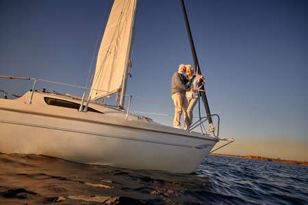 Full length of a romantic retired couple, senior man and woman standing on the side of sail boat or yacht deck floating in the calm blue sea, hugging and enjoying amazing sunsetの写真素材