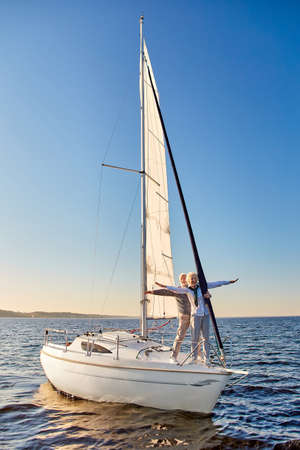 Happy senior couple stretching hands out against the sky, looking at the horizon and enjoying amazing view, standing on the side of sailboat or yacht deck floating in seaの写真素材