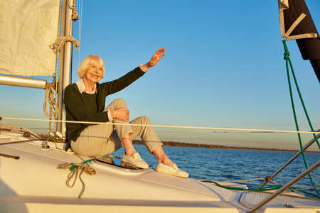 Happy senior woman sitting on the side of sailboat or yacht deck floating in the calm blue sea, looking away and waving to someoneの写真素材
