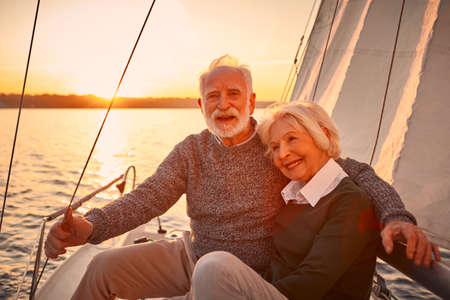 Portrait of a beautiful and happy senior couple in love hugging and enjoying amazing sunset while sitting on the side of sailboat or yacht deck floating in seaの写真素材