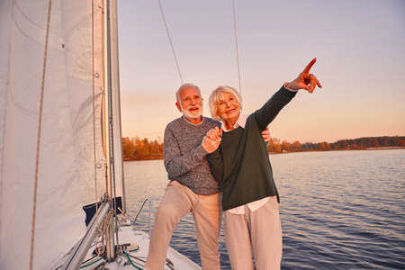 Happy senior couple holding hands and smiling while standing on the side of yacht deck floating in sea, woman pointing at the horizon, sailing at sunsetの写真素材