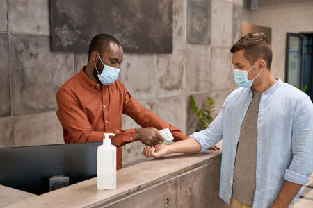 Young african man wearing medical mask standing at reception in the modern office and measuring body temperature of a caucasian male office worker with infrared thermometerの写真素材
