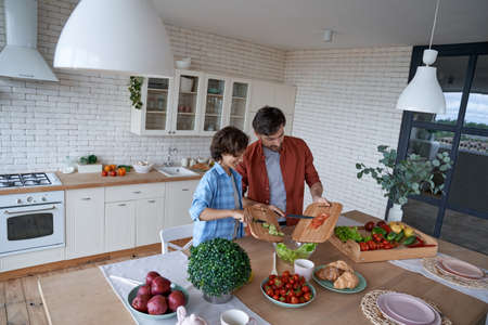 Cooking lunch with dad. Young father and his happy son preparing a salad while standing in the modern kitchen at home, spending time togetherの写真素材