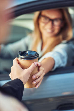 Coffee to go, happy caucasian businesswoman sitting behind steering wheel of her modern car and buying take away coffee, focus on the paper cupの写真素材