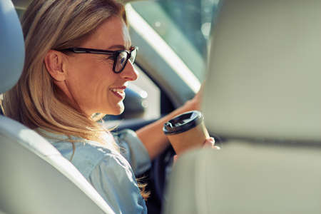 Rear view of a beautiful middle aged business woman wearing eyeglasses sitting behind steering wheel in a car, drinking coffee and smilingの写真素材