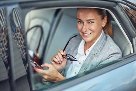 Portrait of a happy mature business woman sitting on back seat in the car and using smartphone, she is looking at camera and smilingの写真素材