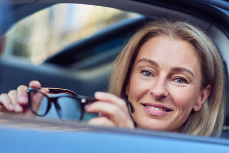 Portrait of a happy business woman holding eyeglasses and smiling at camera while sitting behind steering wheel of her modern car, driving through the cityの写真素材