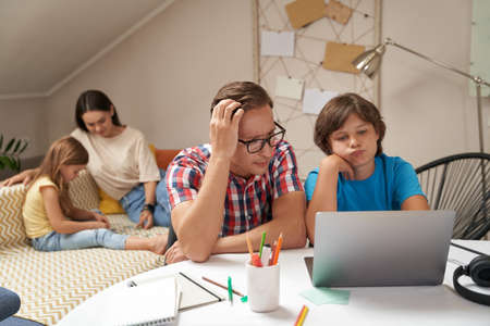 Worried father helping his little son with homework while mother and daughter sitting on sofa and playing with pazzlesの写真素材