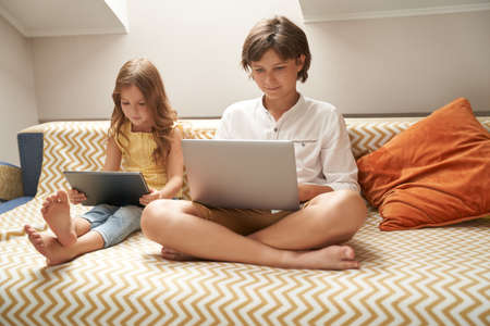 Little children, cute brother and sister sitting on sofa and using laptop and digital tablet, watching cartoons or playing games while spending time together at homeの写真素材