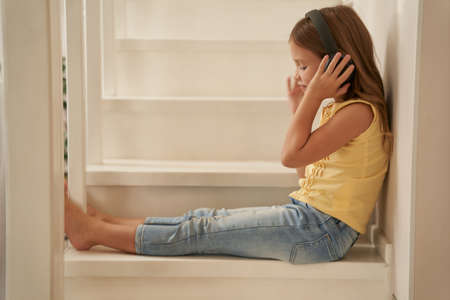 Cute little girl wearing headphones sitting on stairs at home and listening musicの写真素材
