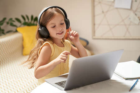 Cute little caucasian girl in headphones using laptop while sitting at the table in her room, watching video lesson at homeの写真素材